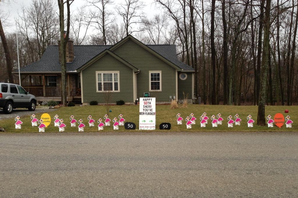 Green ranch home with row of flamingo yard signs for Sheri's 50th birthday