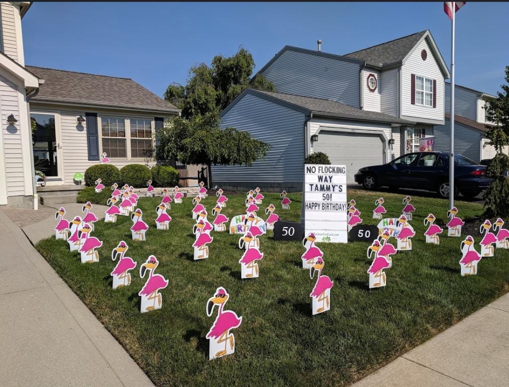 Gray suburban home with large flock of cartoon flamingo yard signs for Tammy's 50th birthday